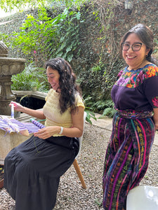 Student weaving on a backstrap loom, and teacher observing, both with a big smile.
