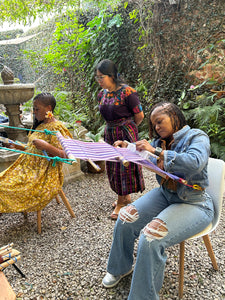 Teacher guiding two students on backstrap looms in front of a founrtain
