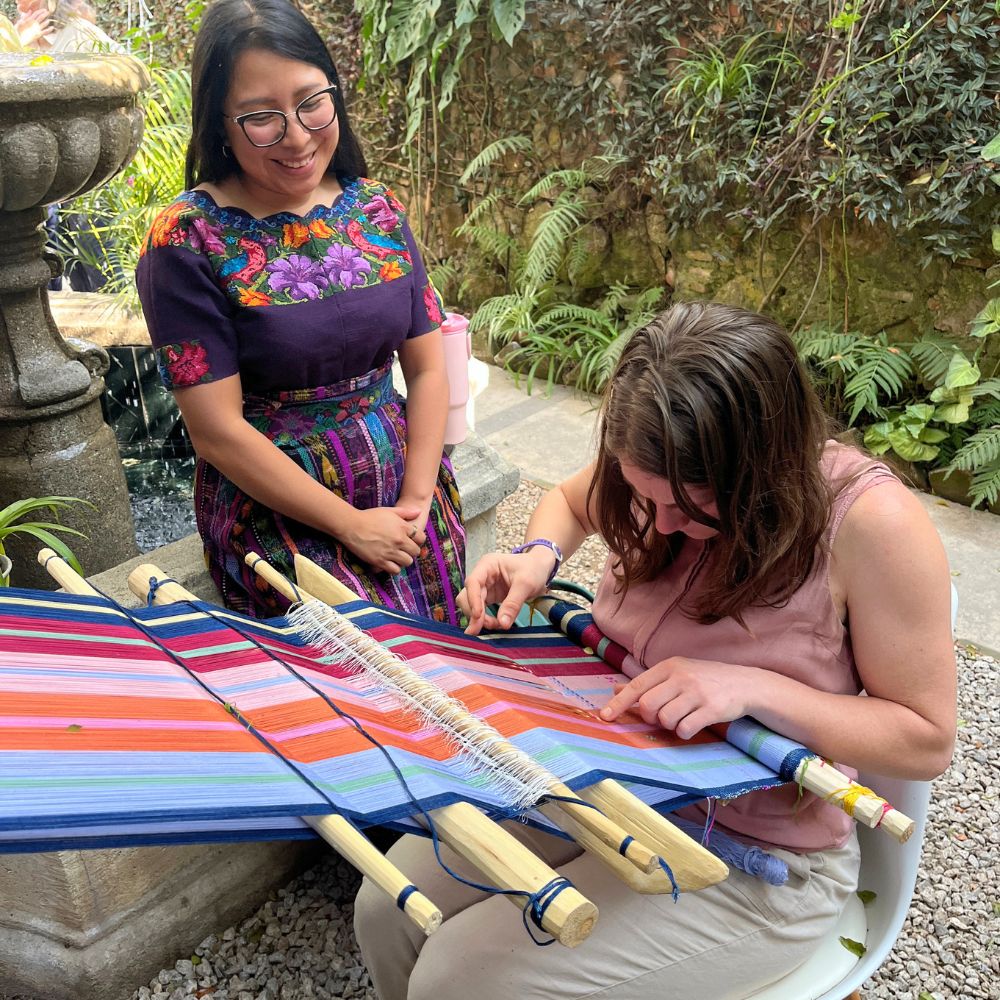 A weaving teacher smiling as she observes her student’s progress on a colorful backstrap loom in the garden beside a relaxing water fountain.