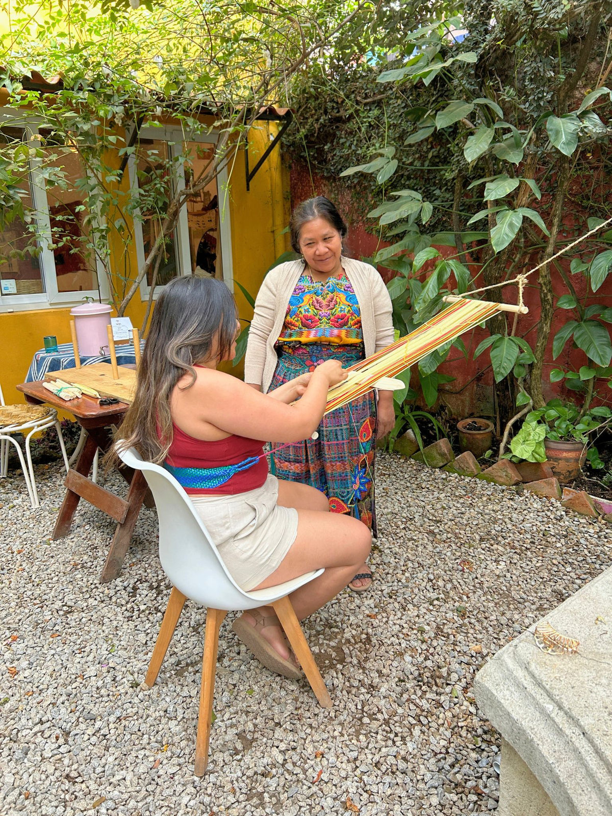 A master weaver and student weaving in the garden on a backstrap loom the student created herself, featuring earth tones and warm orange threads.