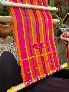 A backstrap loom featuring brocade designs that the student began creating with the guidance of her teacher, using vibrant red threads.