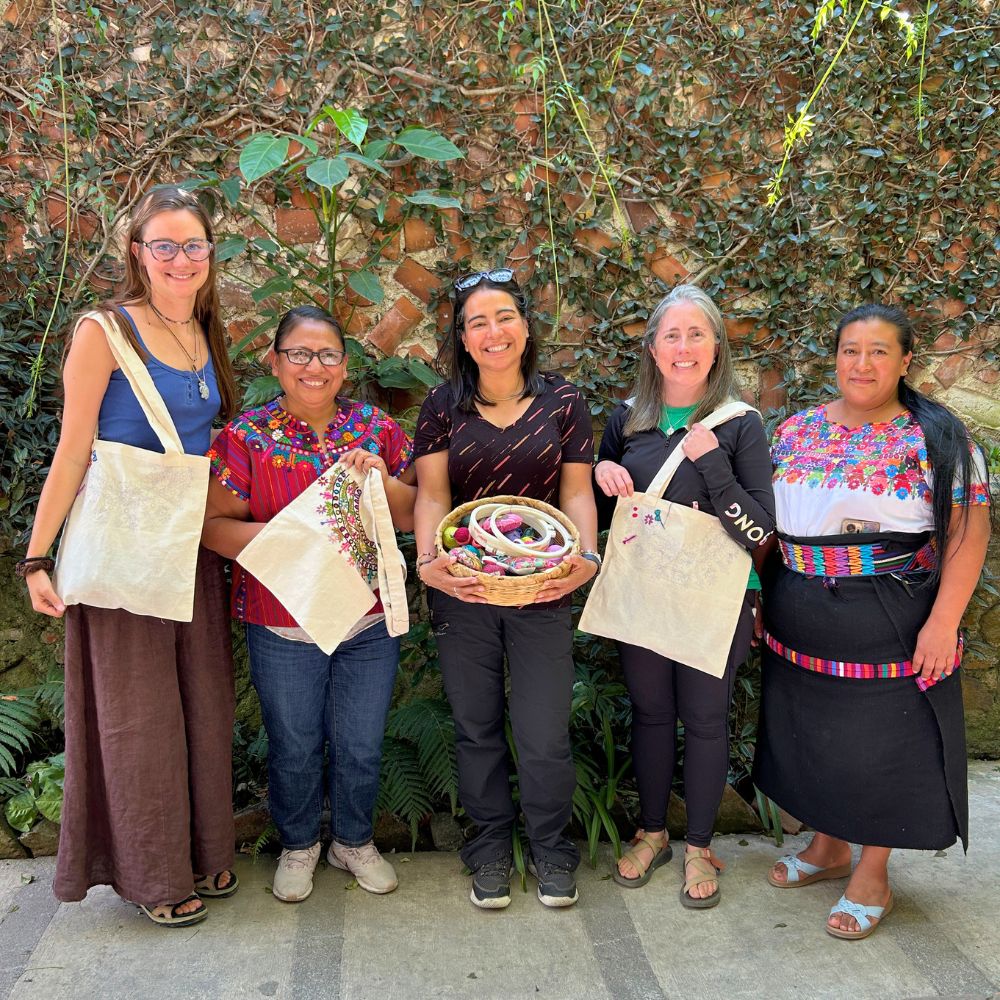 A group of students posing and smiling with their embroidery teachers while displaying their handmade embroidered tote bags.