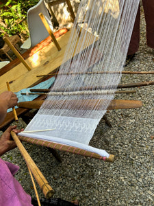 A backstrap loom prepared with white threads for the traditional Pikbil weaving technique from Cobán, showing both the completed woven section and the portion still in progress.