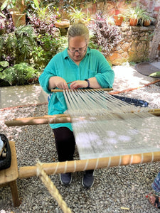 Person weaving with a loom outdoors surrounded by plants