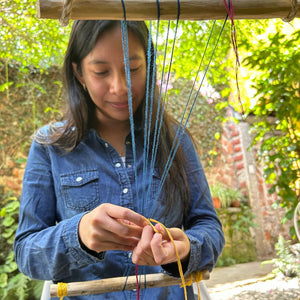 Person weaving with colorful strings using a wooden frame outdoors.