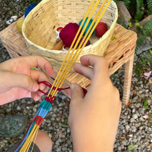 Hands weaving colorful strings on a loom with a basket of yarn in the background.