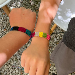 Two children's hands wearing colorful beaded bracelets on a pebbly surface.