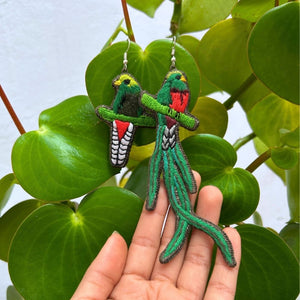 Hand holding quetzal embroidered earrings against a leafy background
