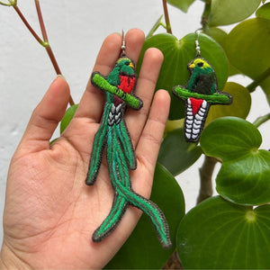 Hand holding embroidered quetzal earrings with a green leafy background