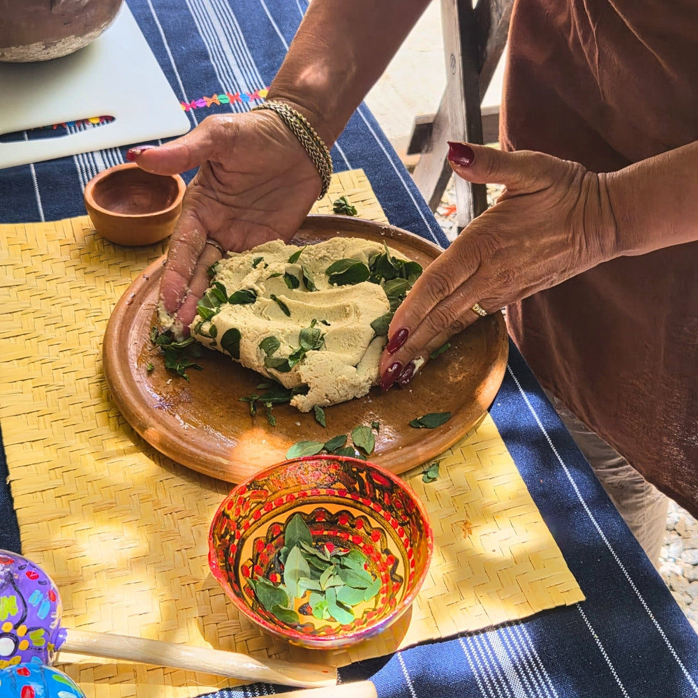 A person's hands are pressing a piece of white corn dough topped with green herbs onto a round wooden serving platter. The scene is set on a blue and white striped tablecloth with a yellow woven placemat, a small clay bowl, and a colorful hand-painted bowl in the foreground.