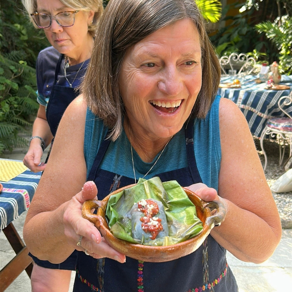 A smiling woman in a blue apron holds a clay bowl filled with green leaf-wrapped food and a red salsa topping in an outdoor garden setting.