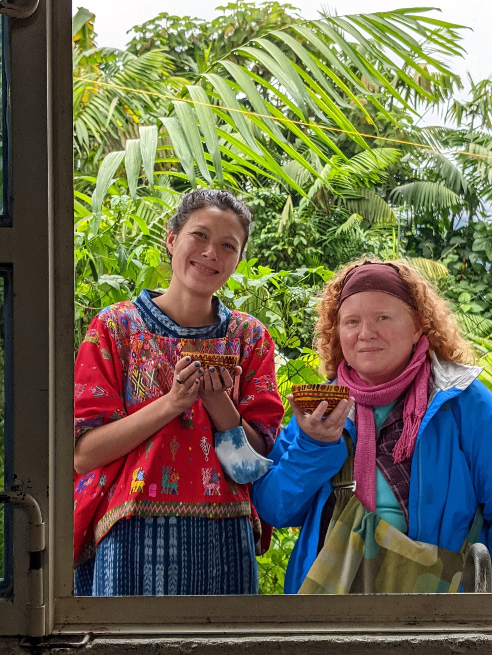 Two women smile through a window, with lush vegetation behind, holding hot cacao gourds