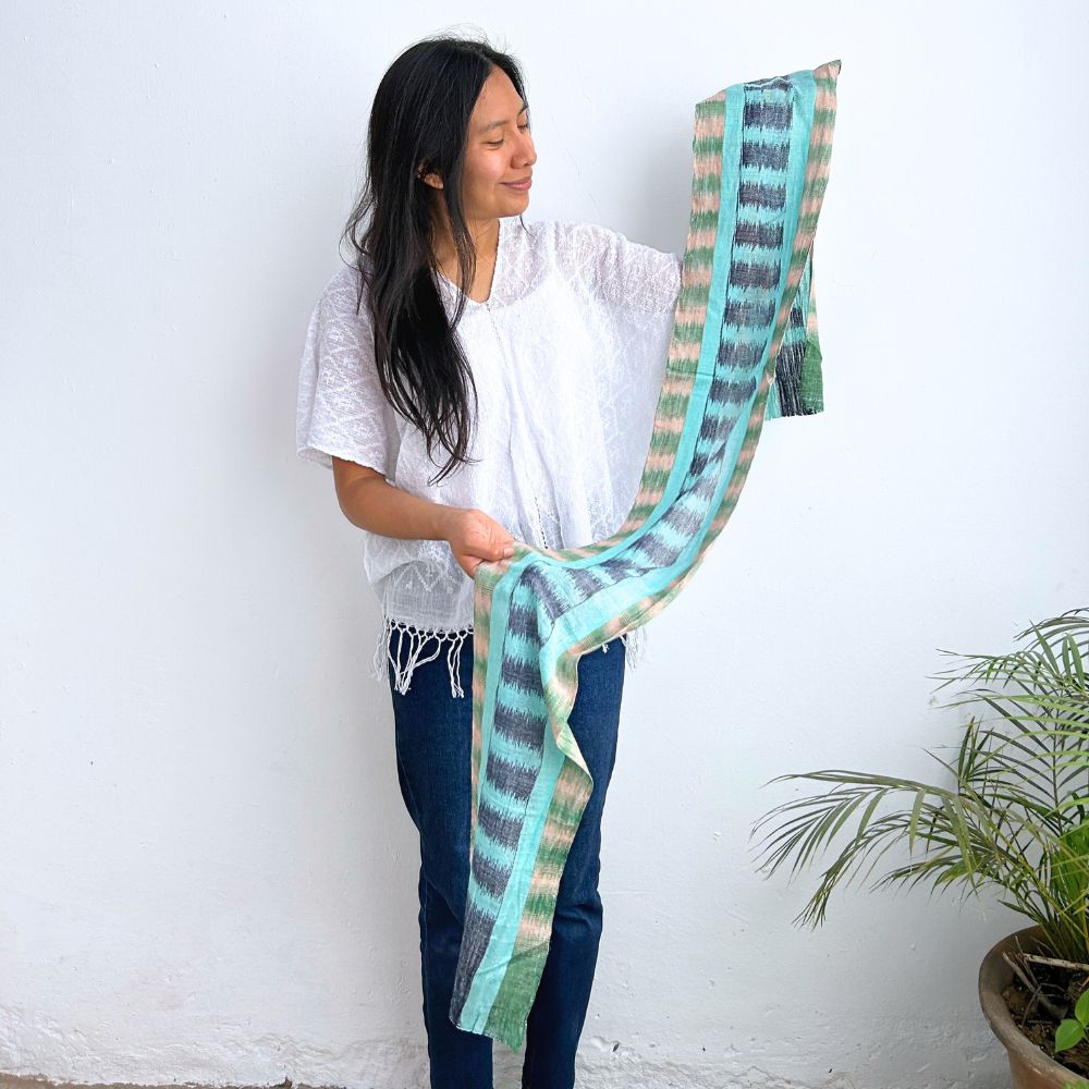 Woman holding a long, patterned scarf against a white wall with a plant in the corner.