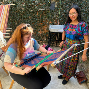 A weaving teacher smiling alongside her student while she weaves on a traditional backstrap loom.