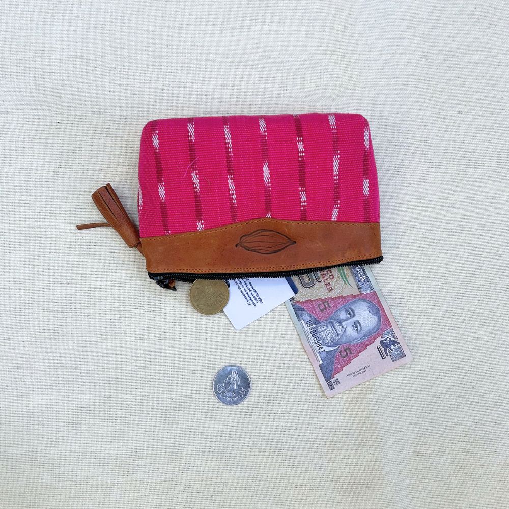 A zippered pouch in bright pink woven fabric with vertical brown stripes and a brown leather trim, shown on a light beige textured fabric background. A coin, a banknote, and a small tag are placed near the pouch.