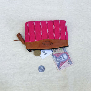 A zippered pouch in bright pink woven fabric with vertical brown stripes and a brown leather trim, shown on a light beige textured fabric background. A coin, a banknote, and a small tag are placed near the pouch.