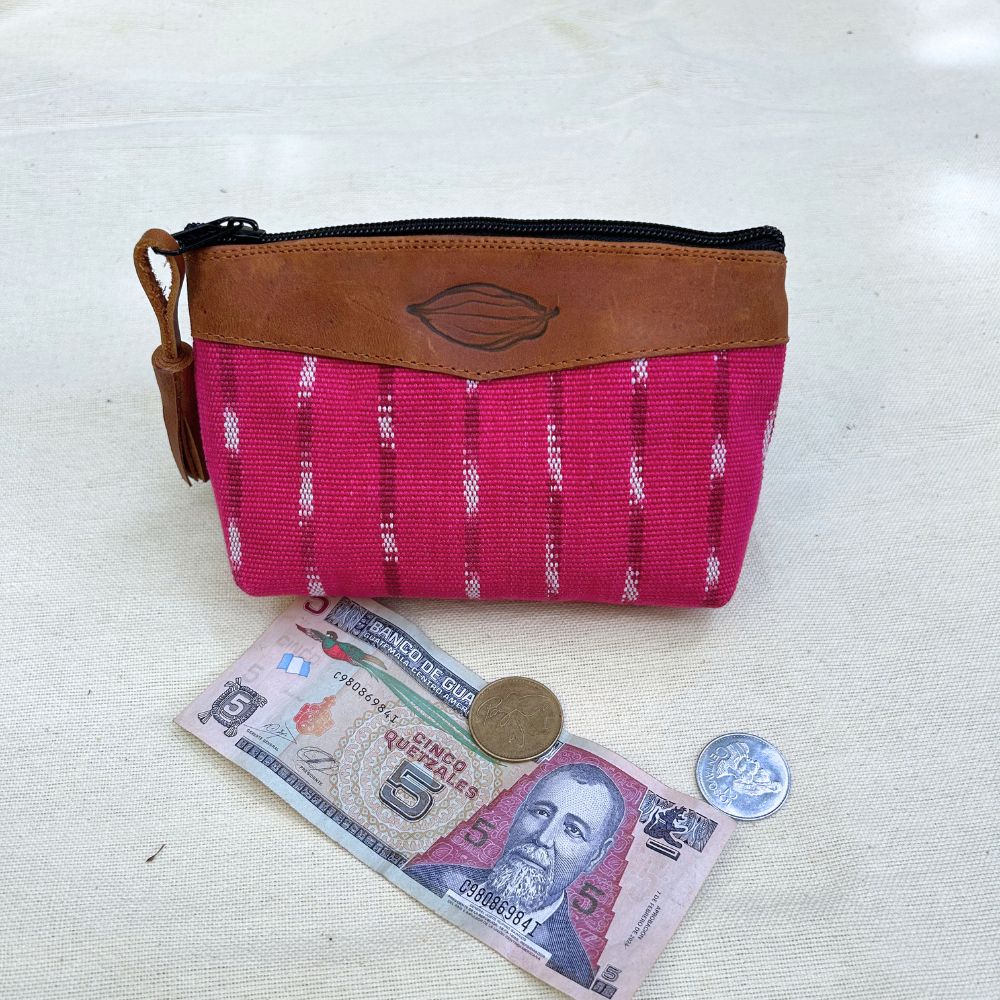 A small zippered pouch with a bright pink patterned fabric body and a brown leather top panel, shown on a light fabric surface with a banknote, coin, and another coin placed in front as scale props.