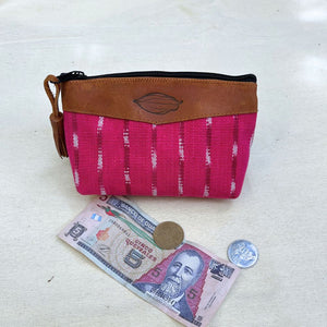 A small zippered pouch with a bright pink patterned fabric body and a brown leather top panel, shown on a light fabric surface with a banknote, coin, and another coin placed in front as scale props.