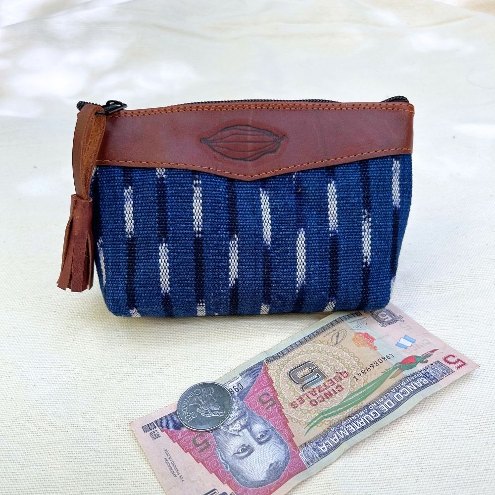 A woven pouch with a blue and white patterned body and brown leather trim, shown on a light fabric surface with a banknote and coin placed in front for scale.