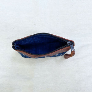 A zippered pouch shown from above on a light beige fabric background. The pouch has a blue and white patterned exterior, a dark blue interior, and a brown leather tassel.