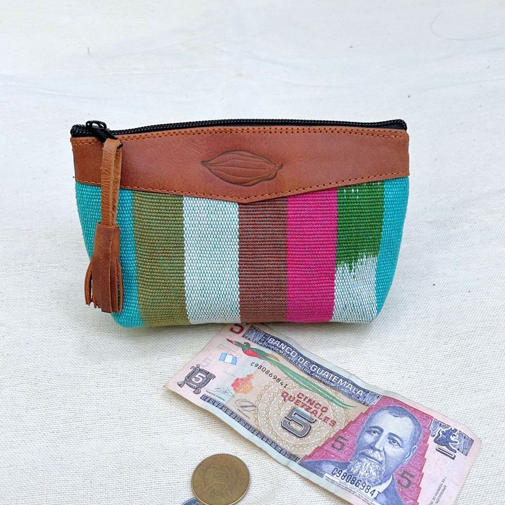 A zippered pouch with a colorful woven striped body and brown leather trim, placed on a light fabric surface with a Guatemalan 5 banknote and a coin below it for scale.