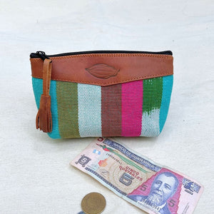 A zippered pouch with a colorful woven striped body and brown leather trim, placed on a light fabric surface with a Guatemalan 5 banknote and a coin below it for scale.