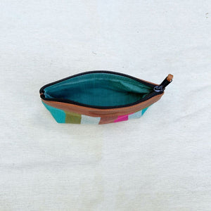 A zippered pouch with a brown leather trim, a teal interior lining, and a multicolor geometric pattern on the exterior, shown open on a light beige fabric background.