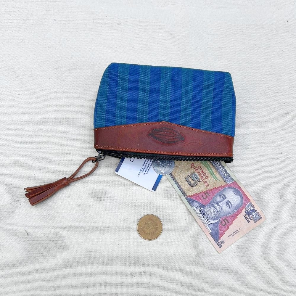 A zippered pouch with a blue and teal striped fabric body and a brown leather trim, shown on a light fabric background with a tassel zipper pull, a banknote, and a coin for scale.