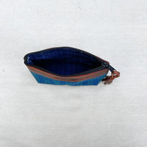 A zippered pouch with a blue and teal patterned fabric exterior, a dark blue interior, and brown leather trim, shown on a light gray textured fabric background.