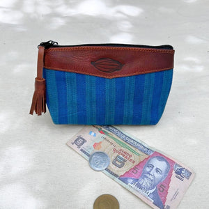 A zippered pouch with a blue and teal striped fabric body and a brown leather top panel, shown on a light fabric surface with a Guatemalan 5 quetzales and coins for scale.