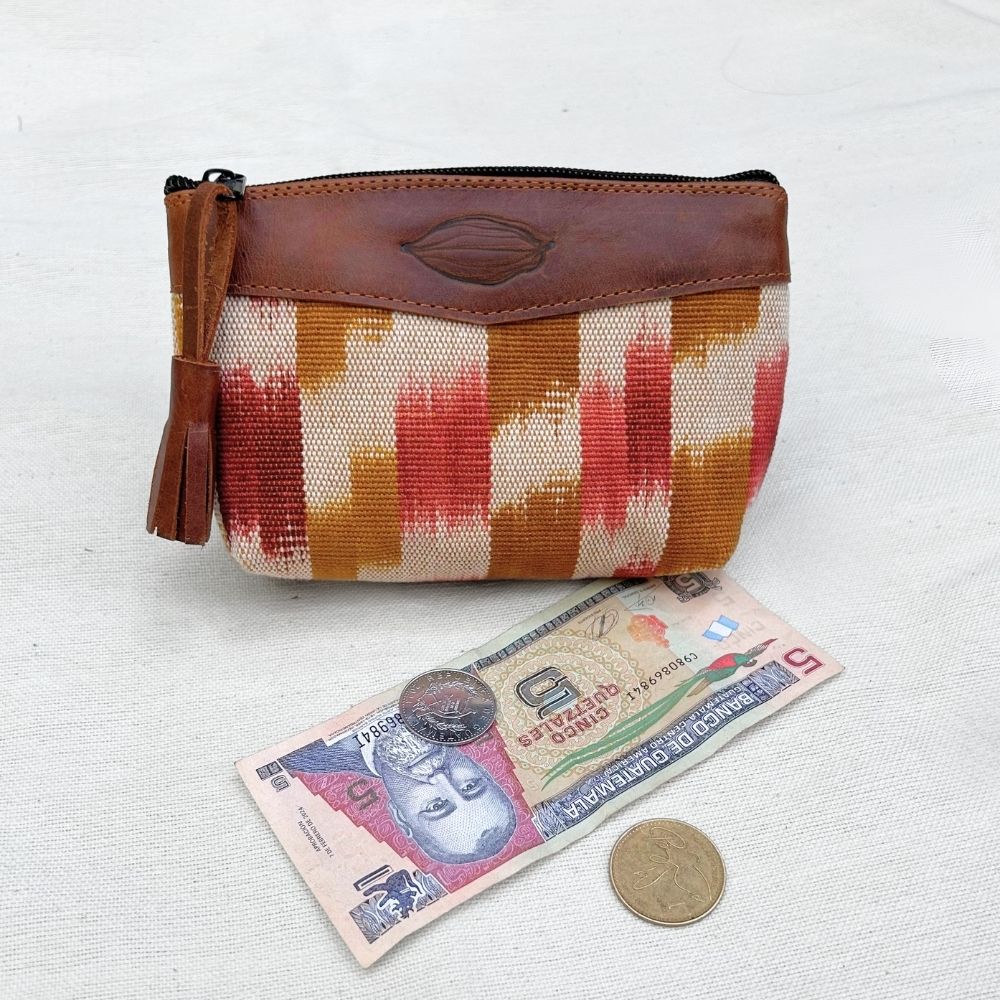 A zippered pouch with a brown leather top panel and a woven body featuring vertical stripes in orange, red, and beige. A tassel hangs from the zipper.