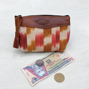 A zippered pouch with a brown leather top panel and a woven body featuring vertical stripes in orange, red, and beige. A tassel hangs from the zipper.