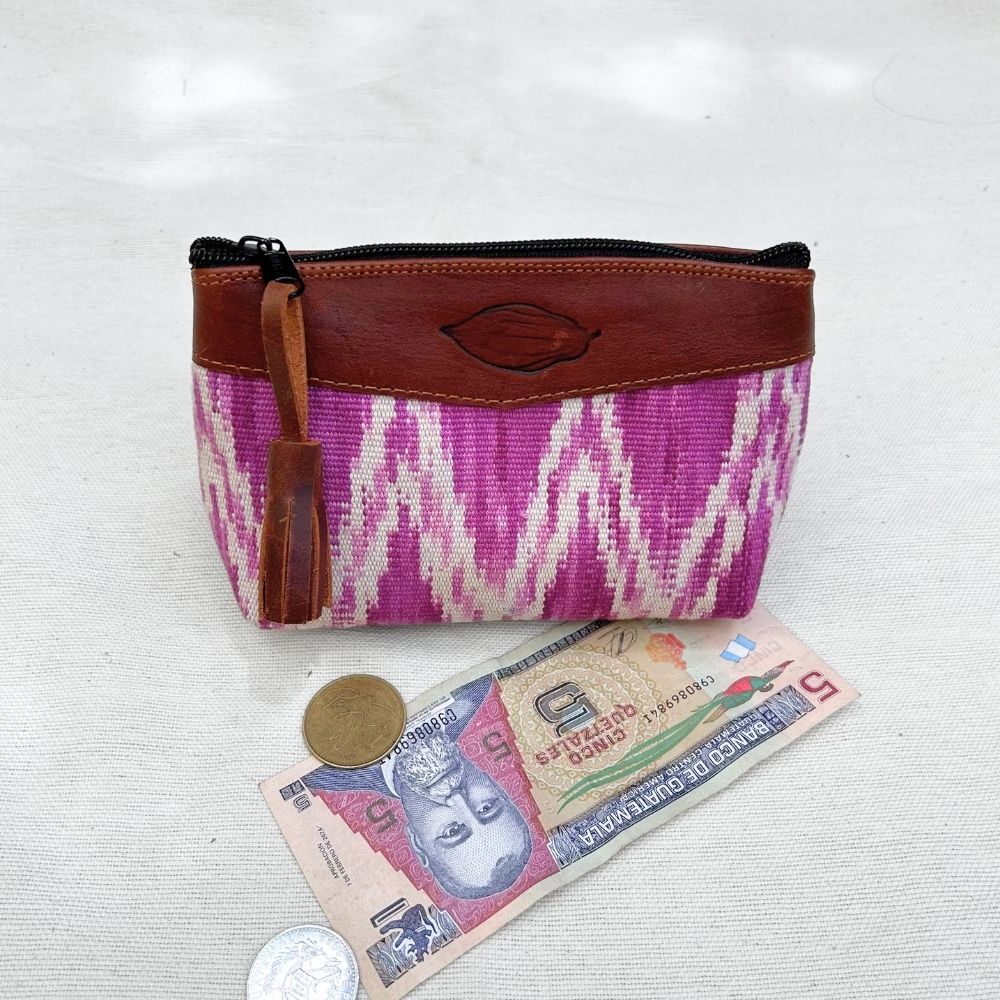 A zippered pouch with a brown leather top panel and a pink-and-white patterned woven body, shown on a light fabric surface with a coin and Guatemalan banknote below for scale.