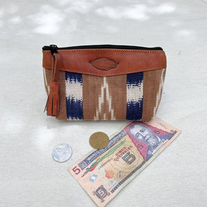 A small zippered pouch with a brown leather top, featuring a woven patterned body in crudo, cream, and blue. The pouch is displayed on a light fabric surface next to a 5 quetzales (Guatemalan bill), a coin, and another coin to indicate its size.