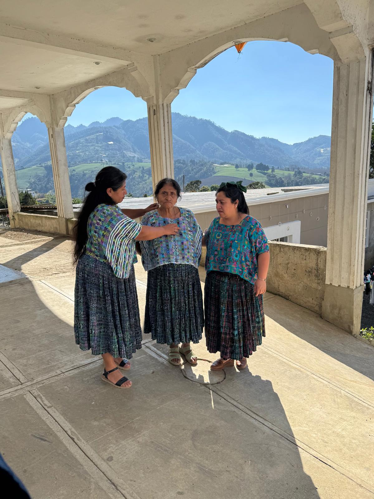 Three women in traditional clothing standing on a balcony with mountains in the background