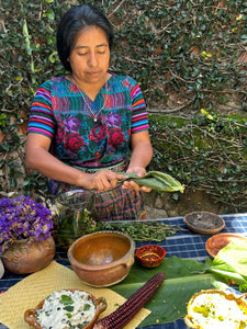 Woman in traditional clothing preparing food outdoors with a table full of ingredients.