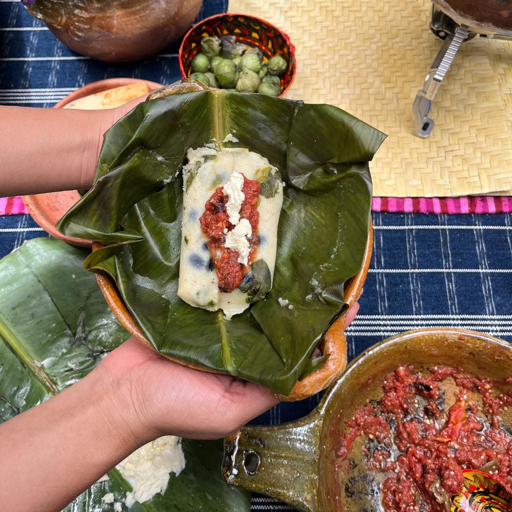 Hand holding a wrapped dish made of leaves with ingredients on a table.