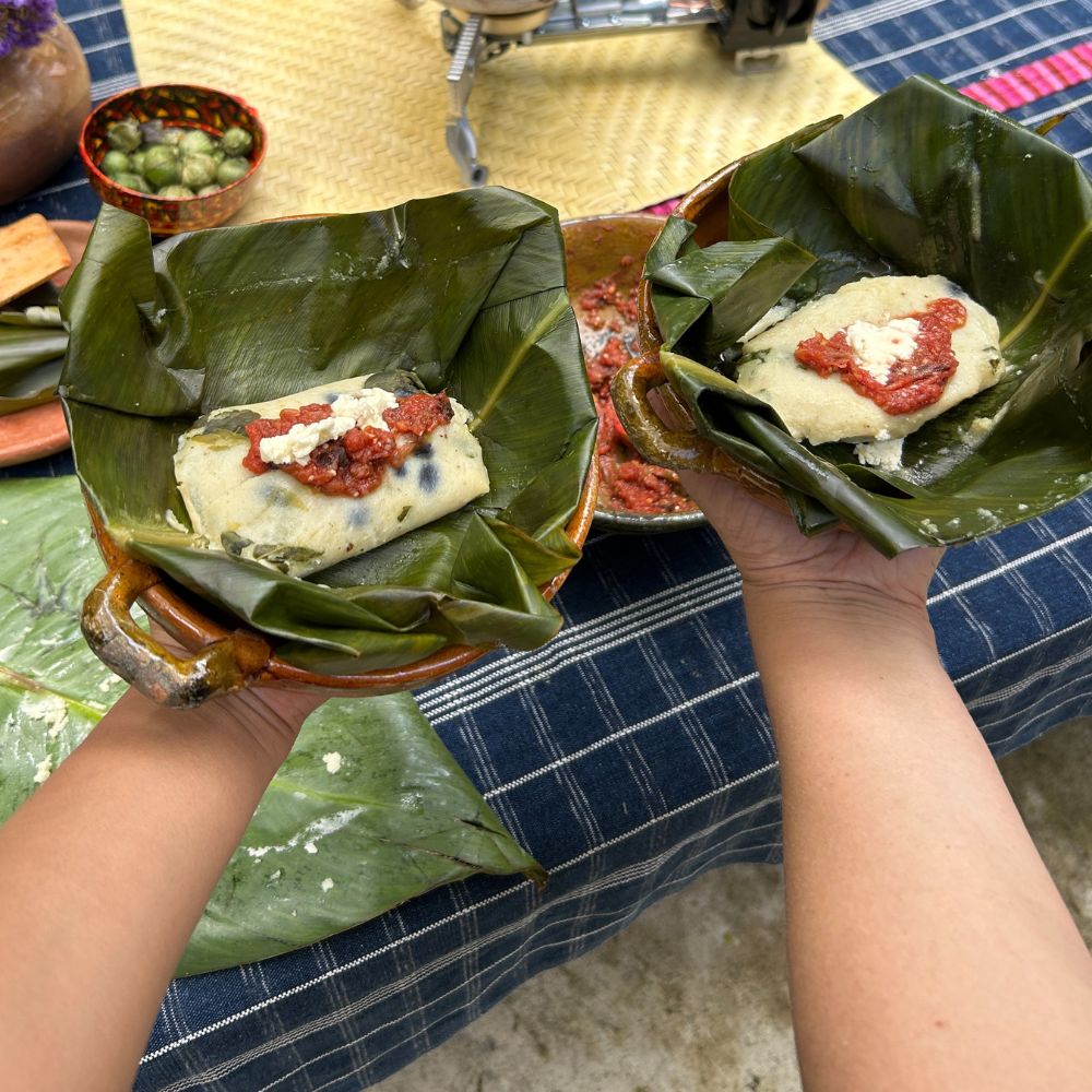 Two hands holding food wrapped in green leaves on a blue checkered tablecloth.