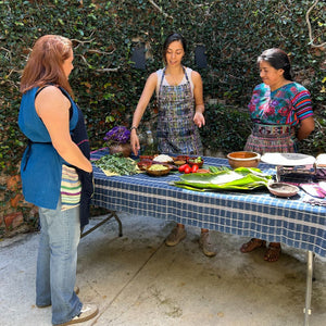 Three women standing around a table with food and cooking ingredients outdoors.