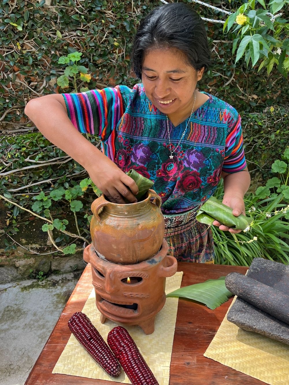 Woman in traditional clothing preparing food outdoors with a wooden table and greenery.
