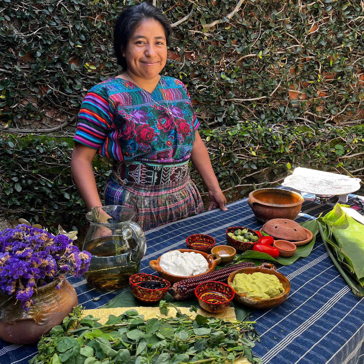 Woman in traditional  Maya clothing standing next to a table with various fresh ingredients for cooking.