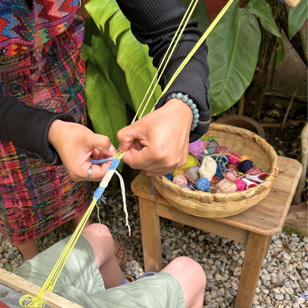 A teacher demonstrating the techniques used to make the bracelet, with a basket of colorful threads in the background for students to choose from.