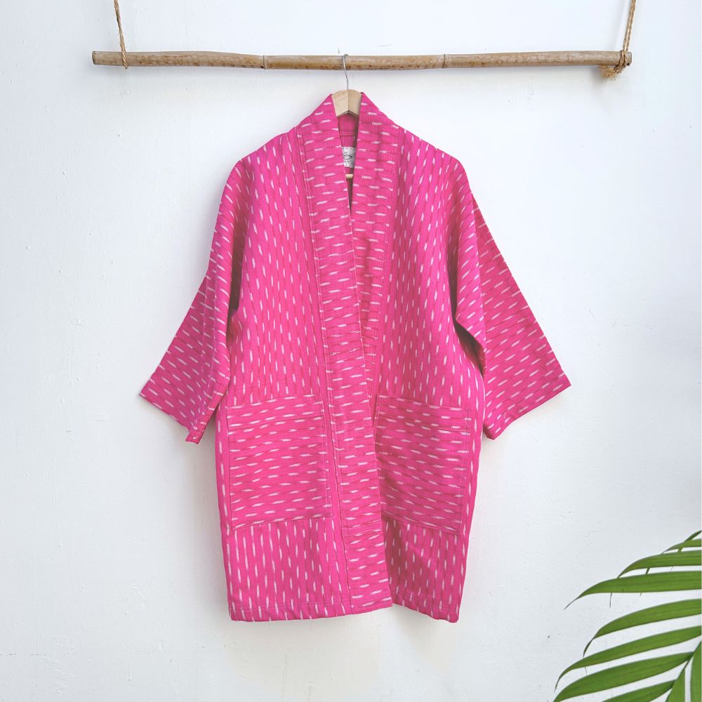 Pink long jacket with white patterns hanging on a wooden rod against a white background.
