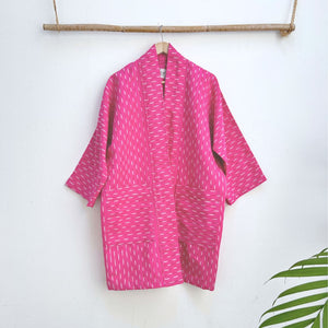 Pink long jacket with white patterns hanging on a wooden rod against a white background.