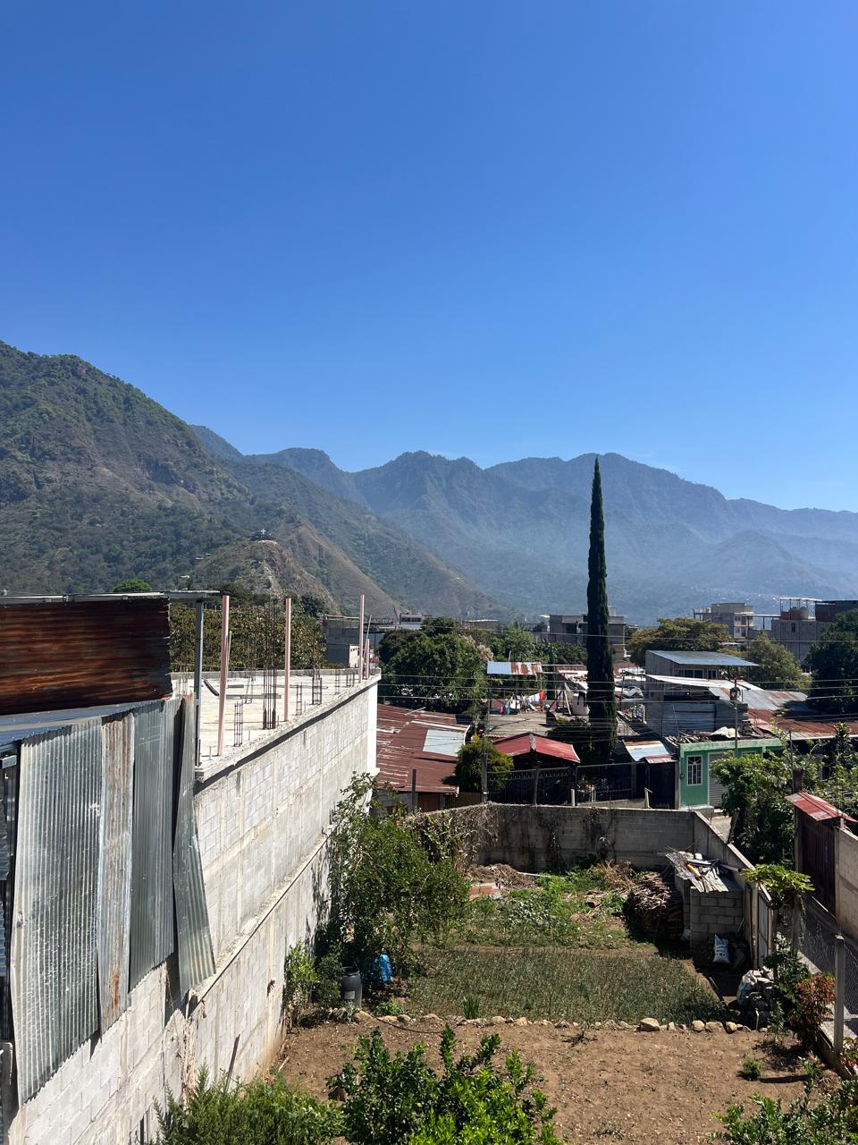 A scenic outdoor view of a mountain town with a tall minaret, rooftops, and green mountains under a clear blue sky, with a concrete wall and small garden plots in the foreground.