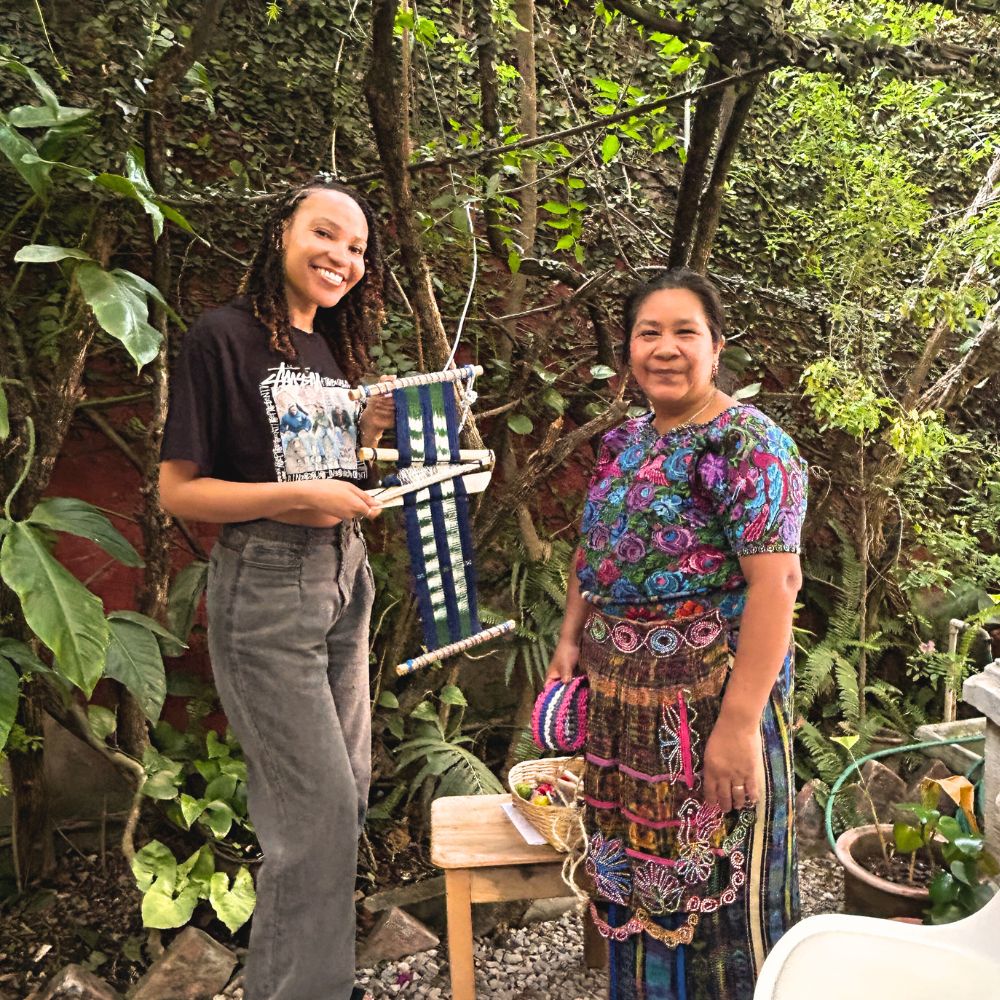 Two women standing outdoors with a loom, surrounded by greenery.
