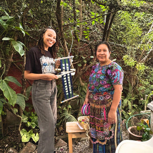 Two women standing outdoors with a loom, surrounded by greenery.