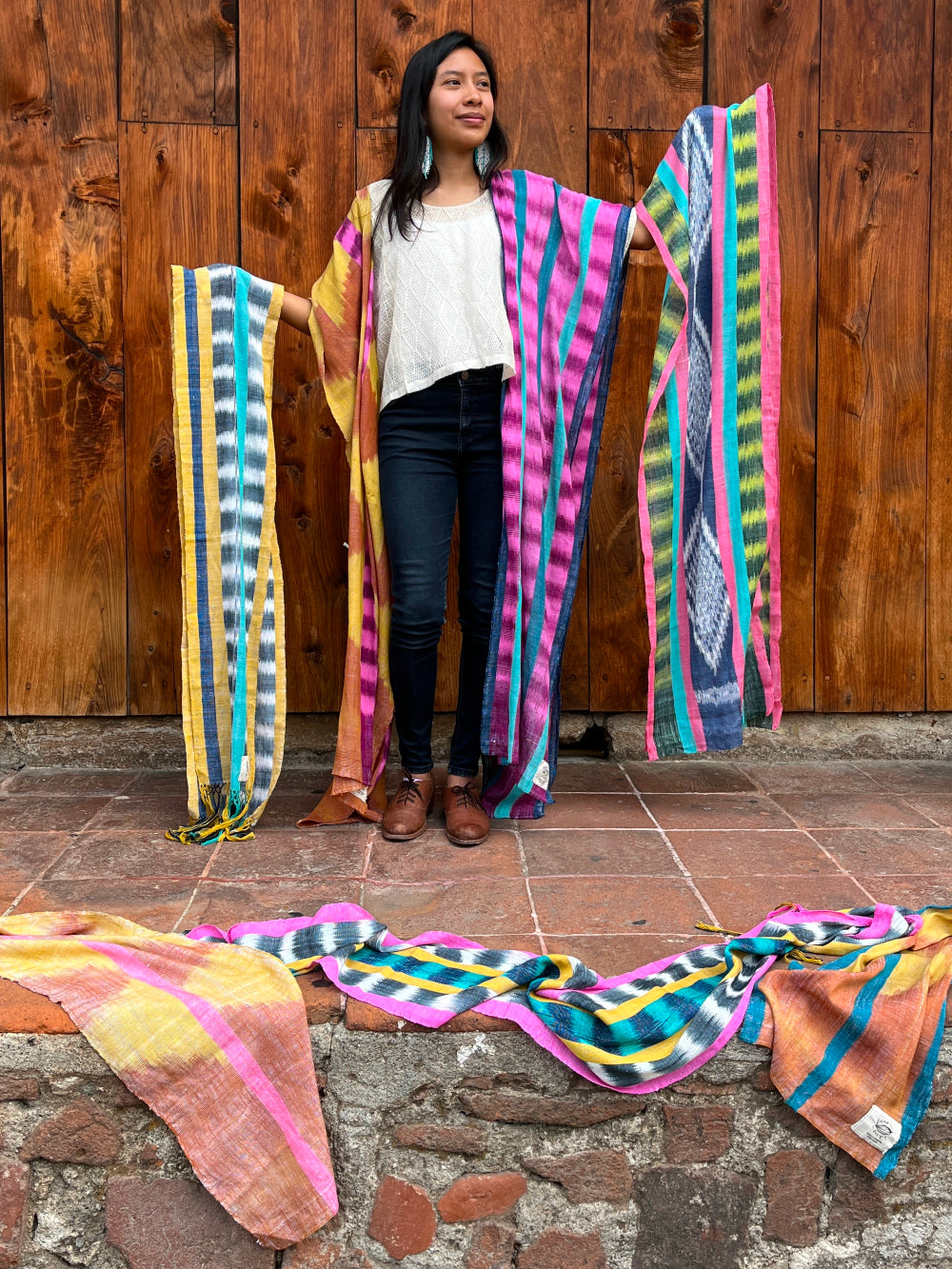 A young woman shows a variety of vibrant handwoven scarves in front of a large wooden door in Antigua Guatemala