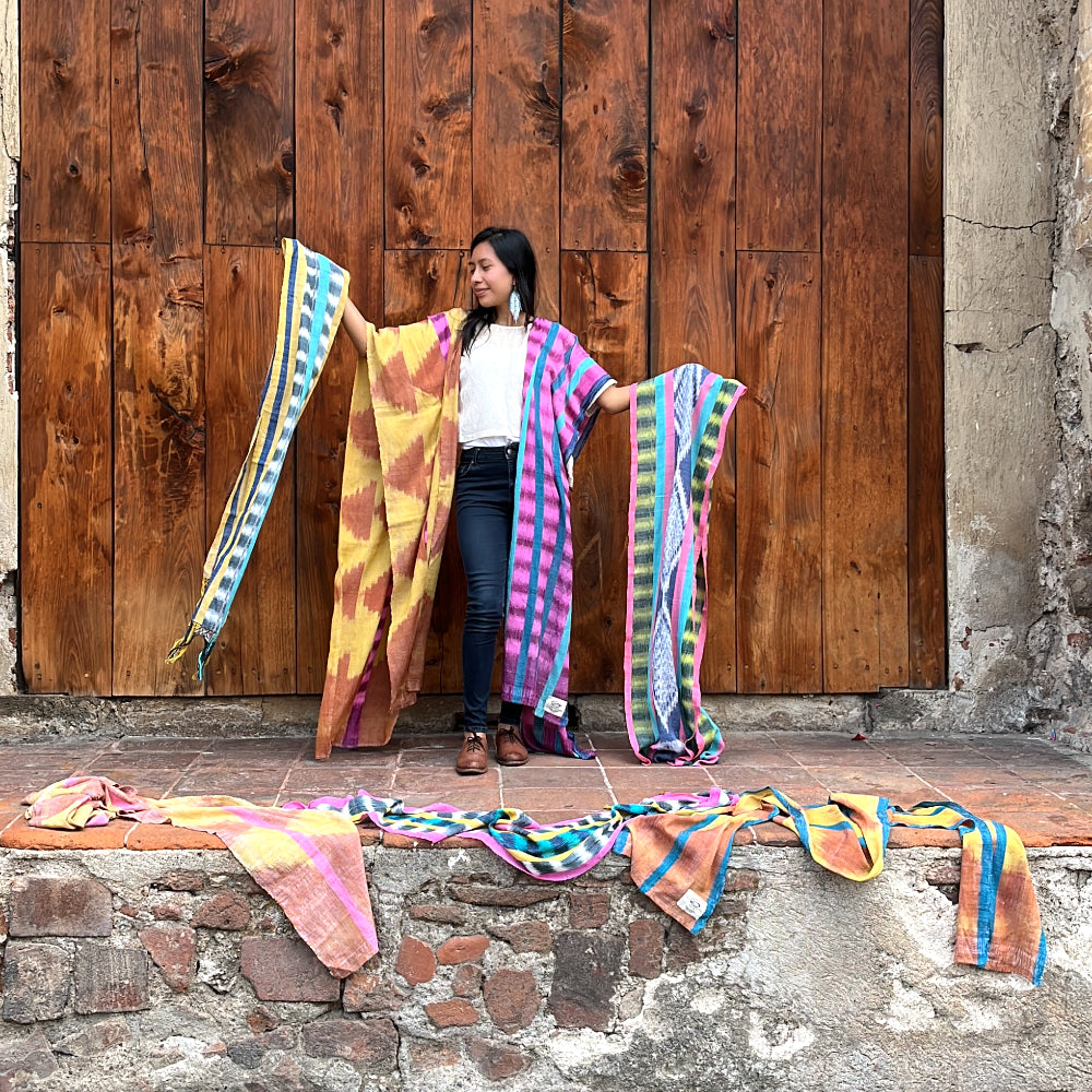 A young woman shows a variety of vibrant handwoven scarves in front of a large wooden door in Antigua Guatemala
