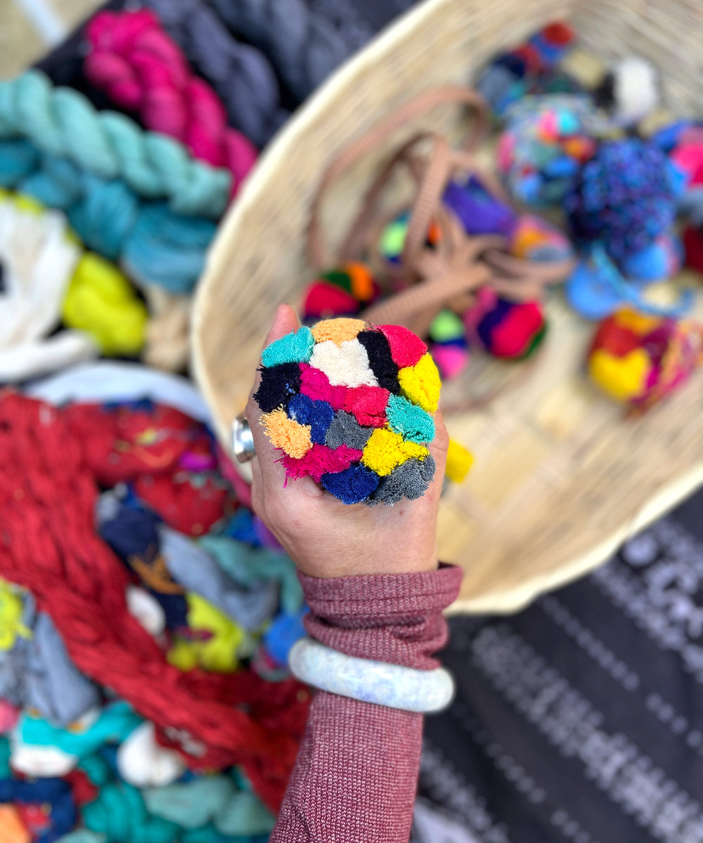 A student's pompom in progress, featuring shades of yellow, pink, white, and gray. In the background, the materials used in the class can be seen, including various naturally dyed cotton yarns.